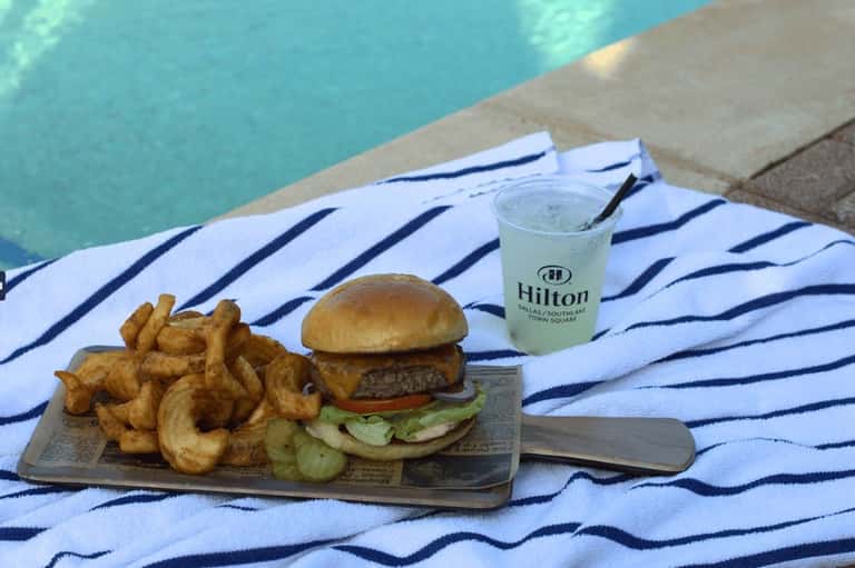 Gourmet burger with curly fries and beverage on striped towel by turquoise pool