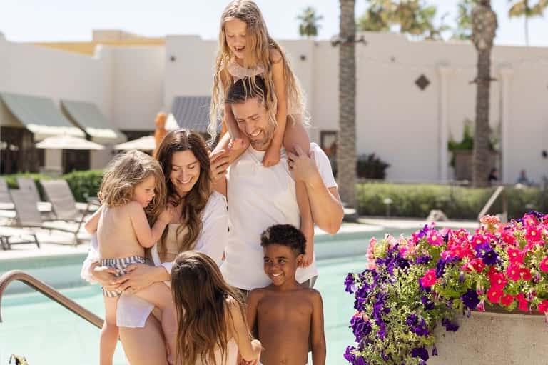Family enjoying pool area with tropical flowers, palm trees, and resort grounds in background