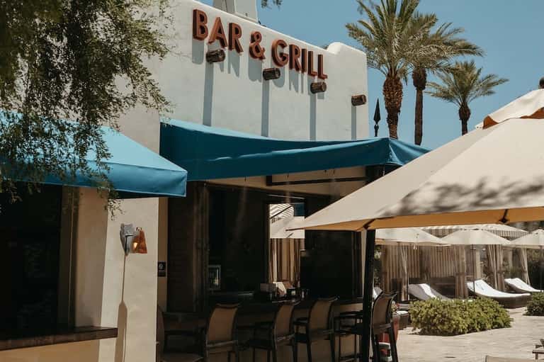 Bar & Grill restaurant exterior with blue awning, palm trees, and patio seating under umbrellas
