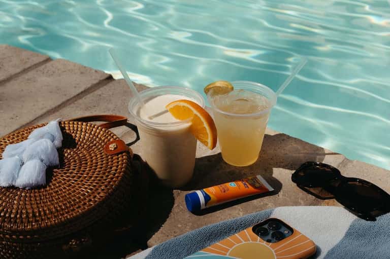 Poolside beverages and sunscreen on concrete deck with turquoise water and sun lounge visible