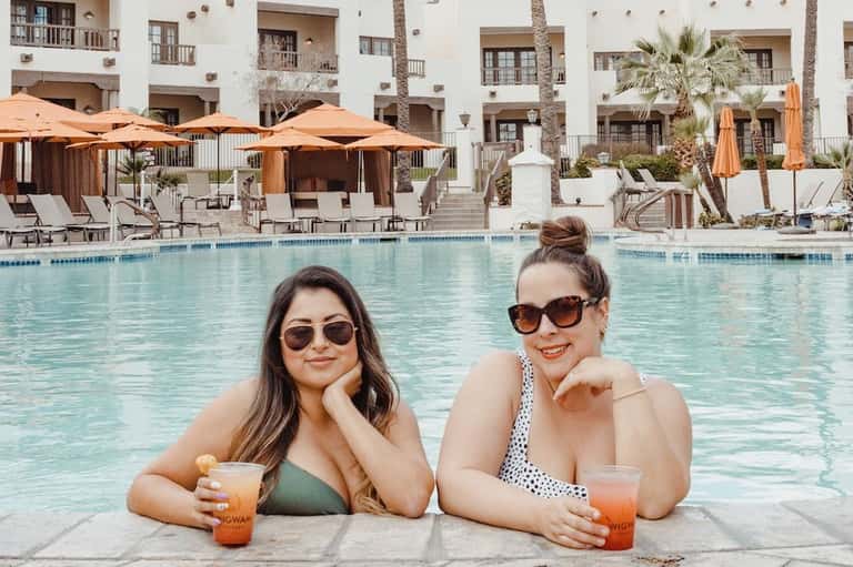 Two women in pool holding drinks with orange umbrellas and white building facade behind