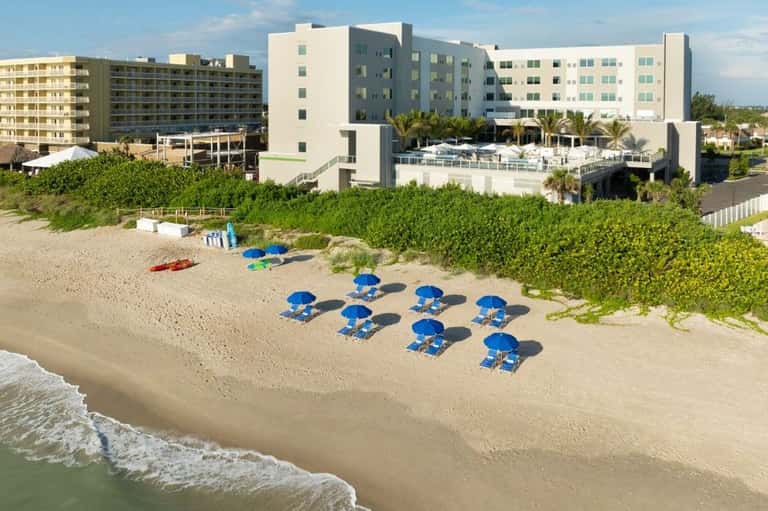 Aerial view of beachfront resort with blue umbrellas, white building, and ocean waves