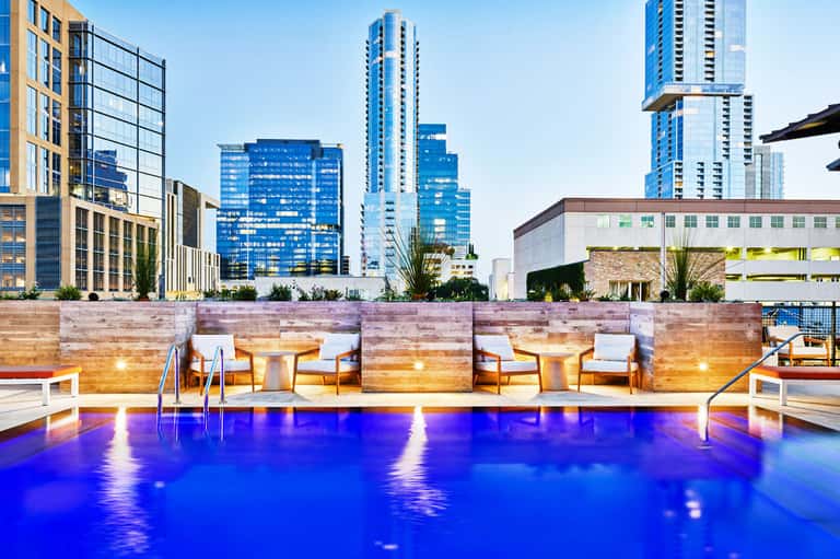Modern rooftop pool with glowing blue water, wooden loungers, and city skyline backdrop