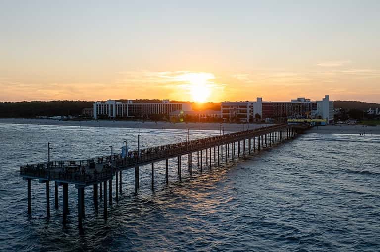 Wooden pier extending into ocean at sunset with beachfront hotels in background