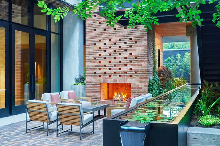 Modern courtyard with brick fireplace, lounge seating, water feature, and hanging greenery