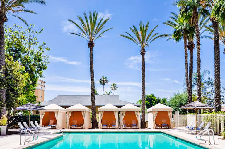 Resort pool with turquoise water surrounded by white cabanas and tall palm trees under blue sky