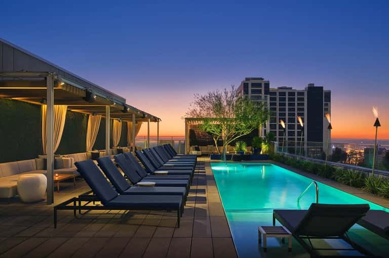 Rooftop pool at sunset with lounge chairs, pergola seating, glowing blue water, and city lights below