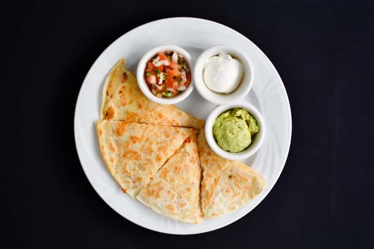Plate of quesadillas with salsa, sour cream, and guacamole served on white dish against black background