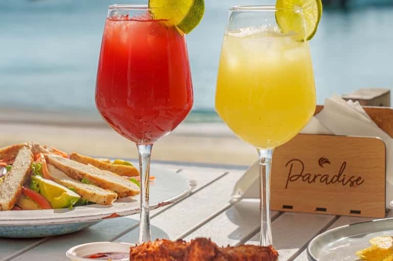 Two colorful tropical cocktails with lime garnish on beach table with Paradise sign