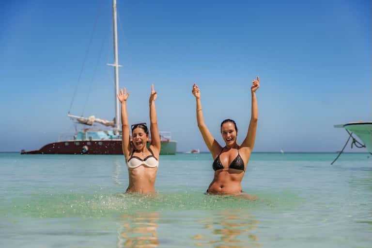 Two women celebrating in turquoise ocean water with sailboat and clear sky