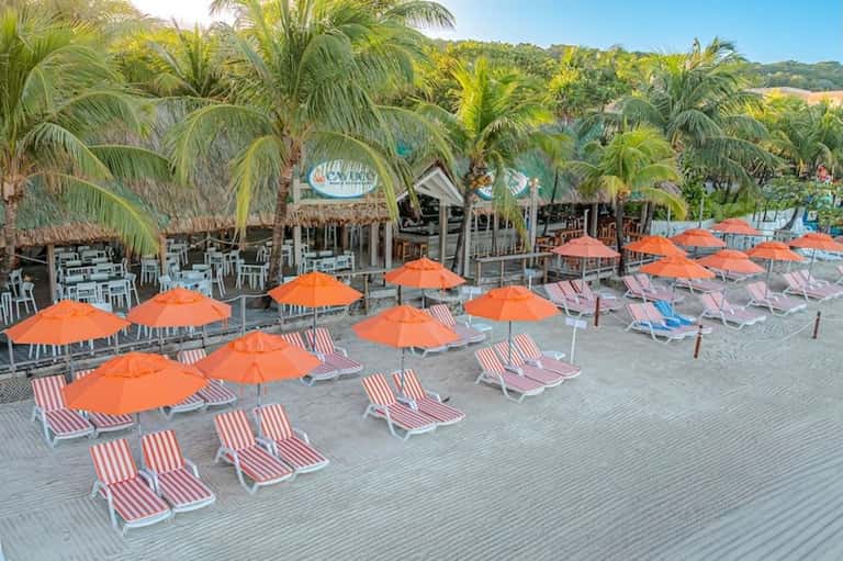 Beachfront deck with orange umbrellas, striped lounge chairs, tiki bar, and palm trees