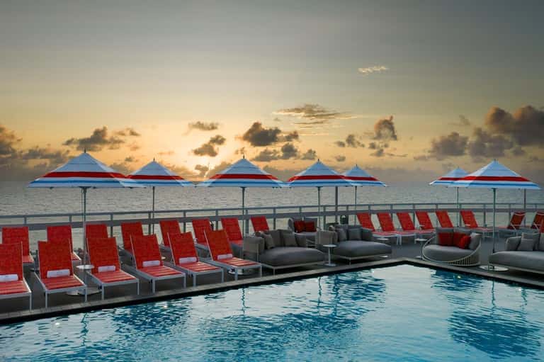 Pool deck at dusk with red lounge chairs under red and blue umbrellas overlooking the ocean at sunset