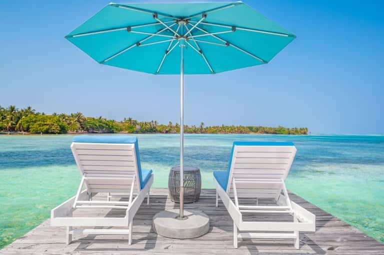 Two white lounge chairs under turquoise umbrella on wooden dock facing pristine tropical lagoon