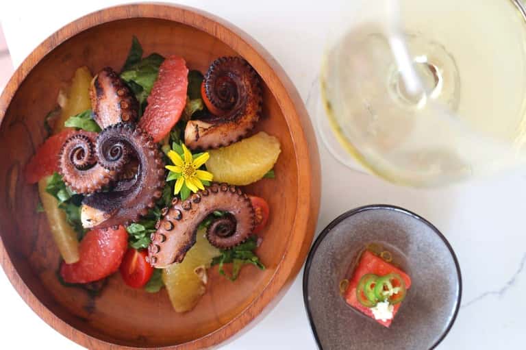 Grilled octopus dish in wooden bowl with wine glass and smaller plated appetizer