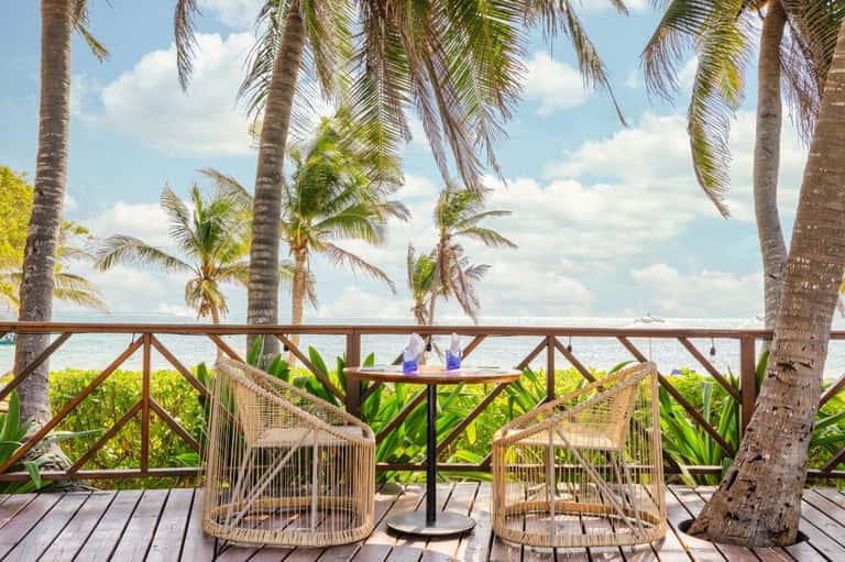 Oceanfront beach seating with woven chairs under palm trees facing crystal blue water