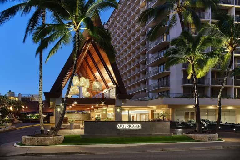 Modern resort entrance at dusk with distinctive A-frame architectural feature, illuminated signage, palm trees, and tall building