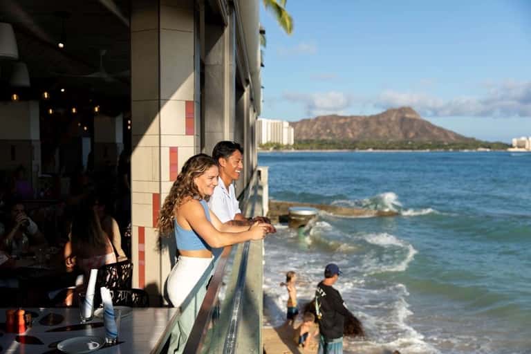 Beachfront bar with couple viewing ocean, Diamond Head mountain, and surfers in water