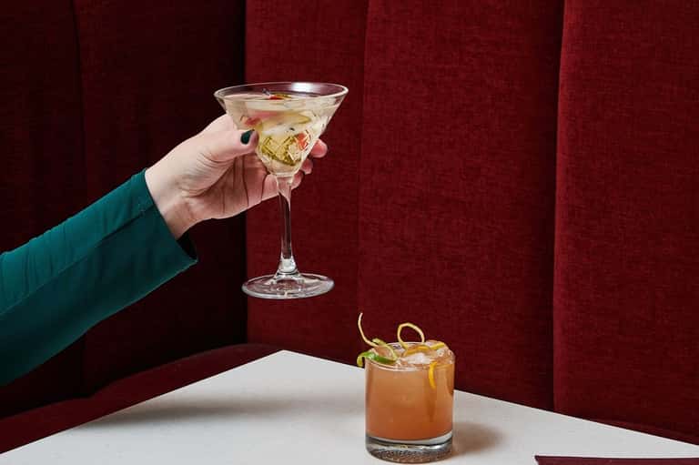 Hand holding a cocktail glass against red velvet curtains with another drink on table
