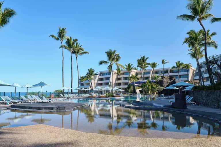 Curved resort pool with shallow area, lounge chairs, umbrellas, and beachfront hotel building