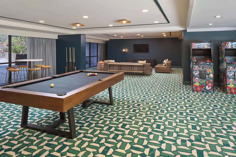 Resort game room with billiards table, arcade games, lounge seating, and geometric patterned carpet