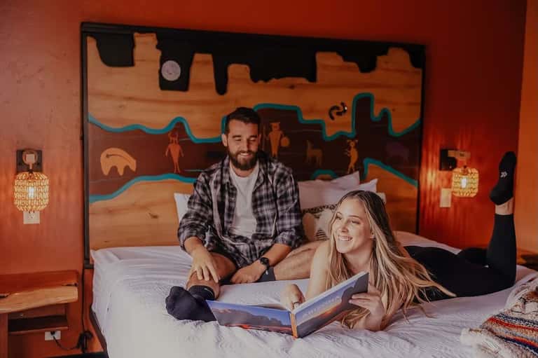 Couple on bed in orange room reading travel magazine with desert artwork headboard