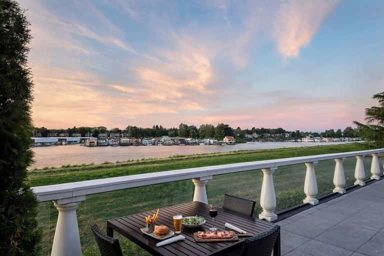 Waterfront balcony dining setup with table, food and drinks, overlooking river at sunset