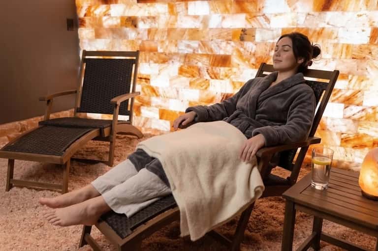 Woman relaxing in salt therapy room with wooden chair, blanket, and glowing salt brick wall