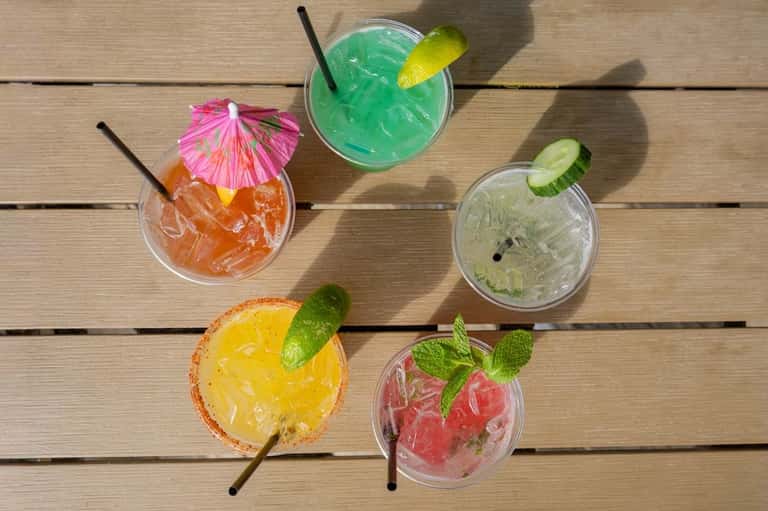 Five artisanal cocktails with fruit garnishes and umbrellas on wooden deck in sunlight