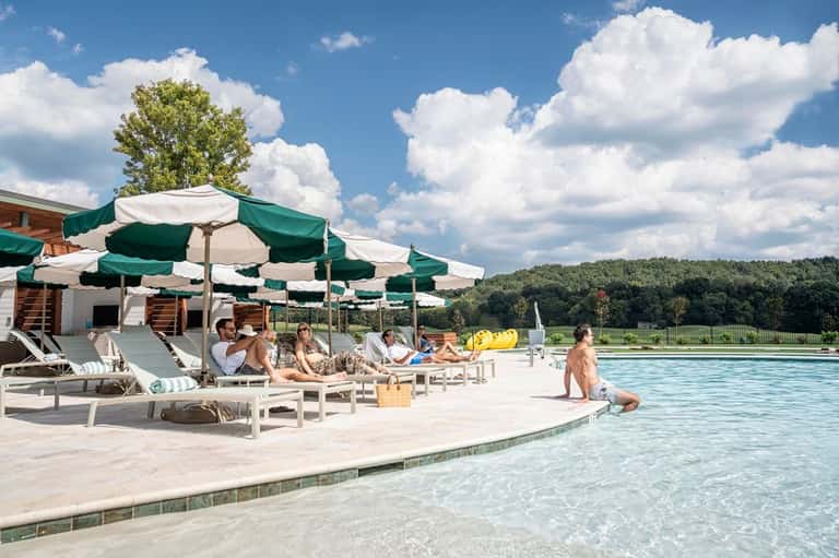 Resort pool area with green and white umbrellas, lounge chairs, sunbathers, and landscaped grounds with trees