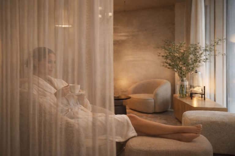 Serene spa lounge with woman holding coffee cup by sheer curtains and curved beige armchair