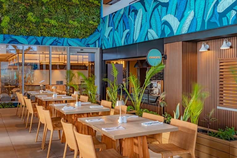 Outdoor restaurant patio with wooden tables, rattan chairs, blue leaf mural, and green living wall