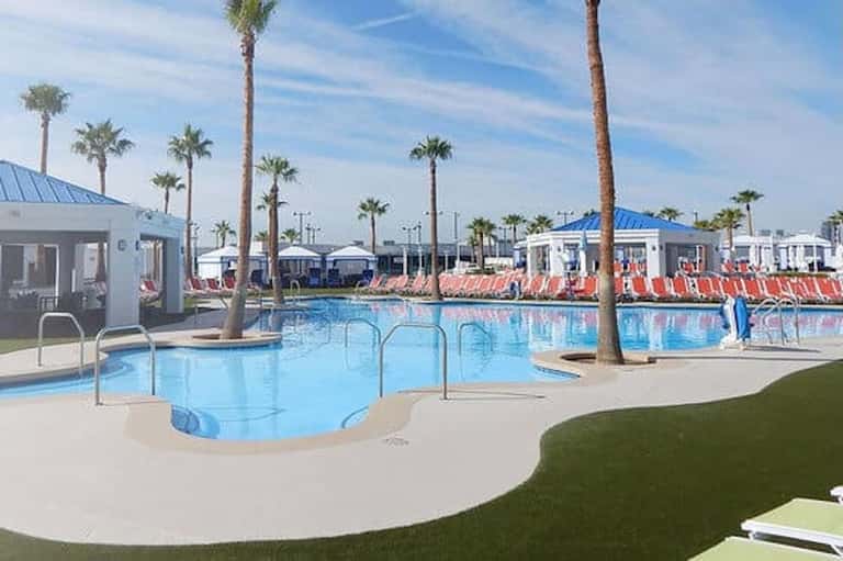 Modern resort pool area with palm trees, red lounge chairs, white buildings, and blue metal roofs
