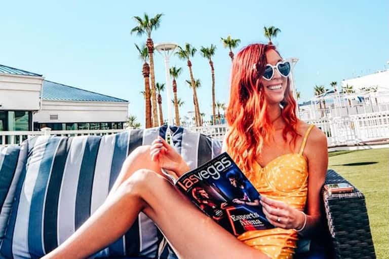 Woman in yellow dress with red hair wearing heart-shaped sunglasses reading magazine by striped lounge chair