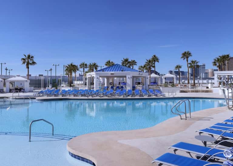 Contemporary resort pool with blue lounge chairs, white pavilion, palm trees, and waterfront setting