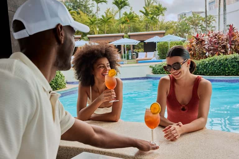 Three guests enjoying cocktails poolside at resort bar with pool in background