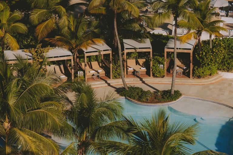Aerial view of wooden cabanas with white canvas tops lining curved resort pool and landscaping