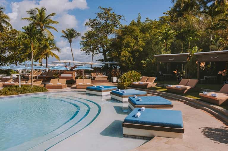 Resort pool with integrated blue lounge chairs, wooden cabanas, ocean view, and tropical landscaping