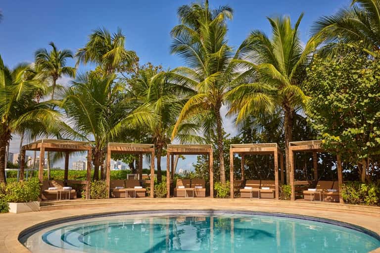 Circular resort pool surrounded by wooden cabanas, palm trees, and manicured green lawn areas