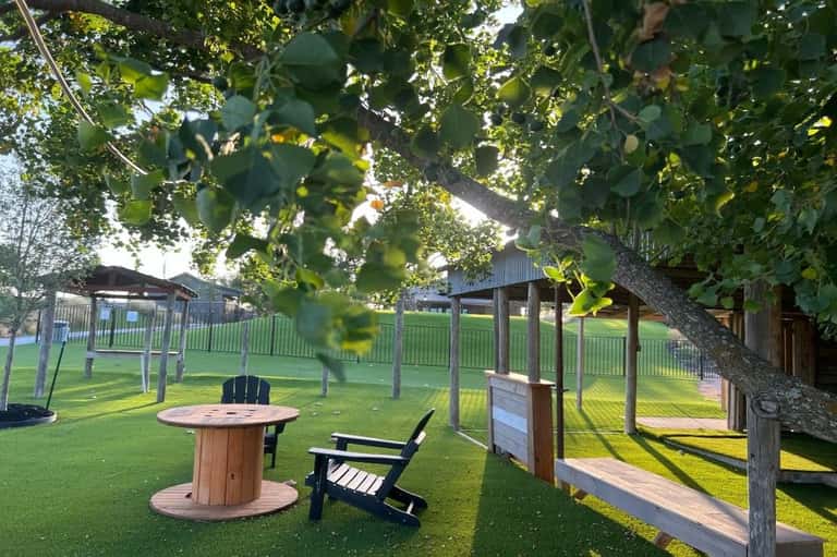 Outdoor seating area with fire pit table, Adirondack chairs, wooden pergolas, and mature trees on green lawn