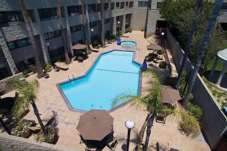 Aerial view of hotel pool courtyard with blue lap pool, surrounding buildings, palm trees, and terrace seating areas