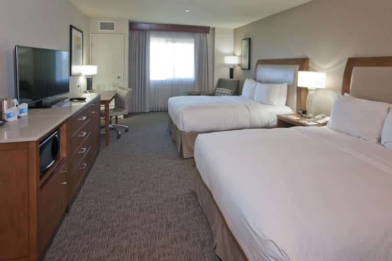 Spacious hotel guest room with two queen beds, dark wood furnishings, flat-screen TV, and large windows