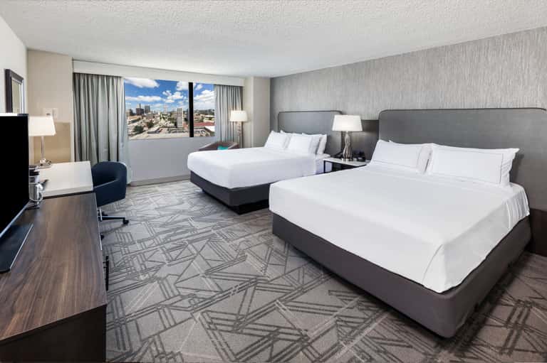Modern hotel room with two queen beds, city skyline view through windows, work desk, and contemporary décor