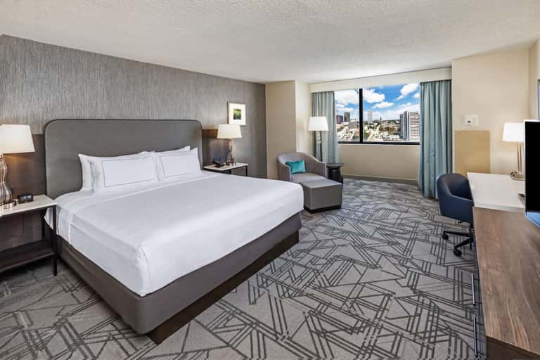 Spacious hotel bedroom with king bed, gray headboard, city view windows, armchair, and geometric patterned carpet