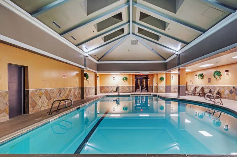 Indoor lap pool with turquoise water, tan tile walls, skylights, and lounge chairs in heated facility