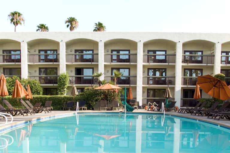 Rectangular pool with orange umbrellas and loungers facing white arched hotel buildings with palm trees