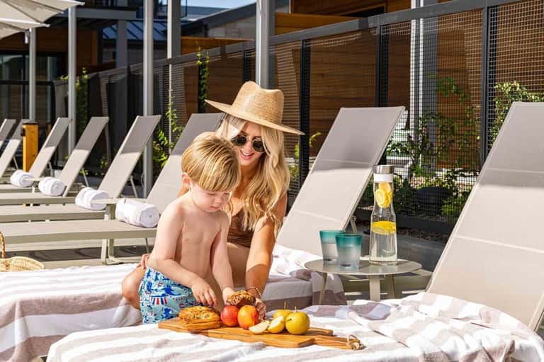 Woman and child enjoying snacks on lounge chairs at a resort pool deck with modern amenities and wooden privacy screens