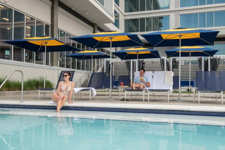 Resort pool with blue and yellow umbrellas, lounge chairs, and modern building facade on sunny day