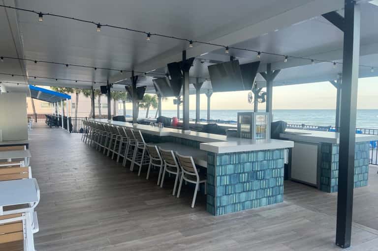 Modern beachfront bar with long counter seating, string lights, and ocean views through open-air covered structure