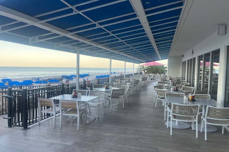 Oceanfront dining patio with white chairs, tables, blue pergola roof, and direct beach access