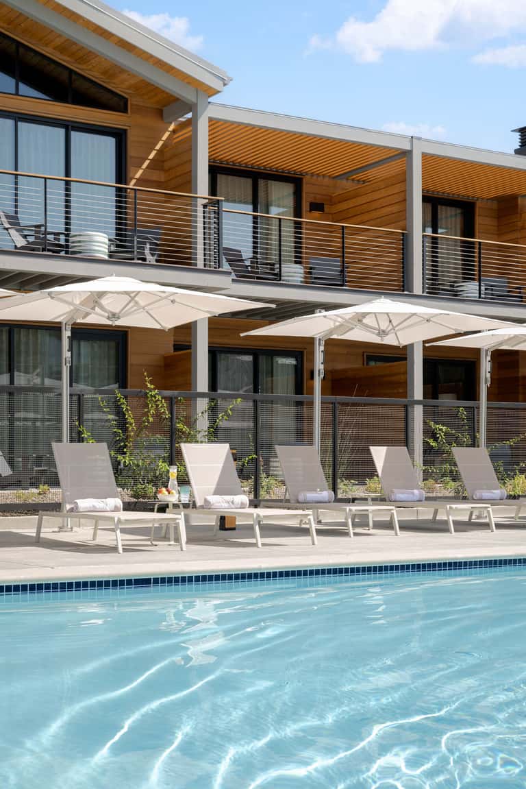 Resort pool area with white umbrellas, lounge chairs, wooden building, and metal railings